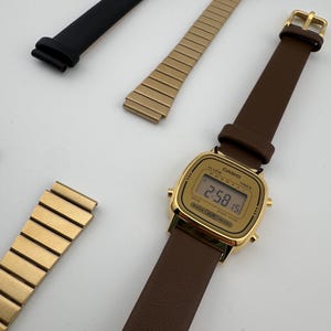 K&ouml;nnte beinhalten: Eine goldfarbene Digitaluhr mit braunem Lederarmband, dazu ein schwarzes Lederarmband und zwei goldfarbene Metallarmb&auml;nder. Das Zifferblatt der Uhr ist rechteckig und zeigt die Uhrzeit an.