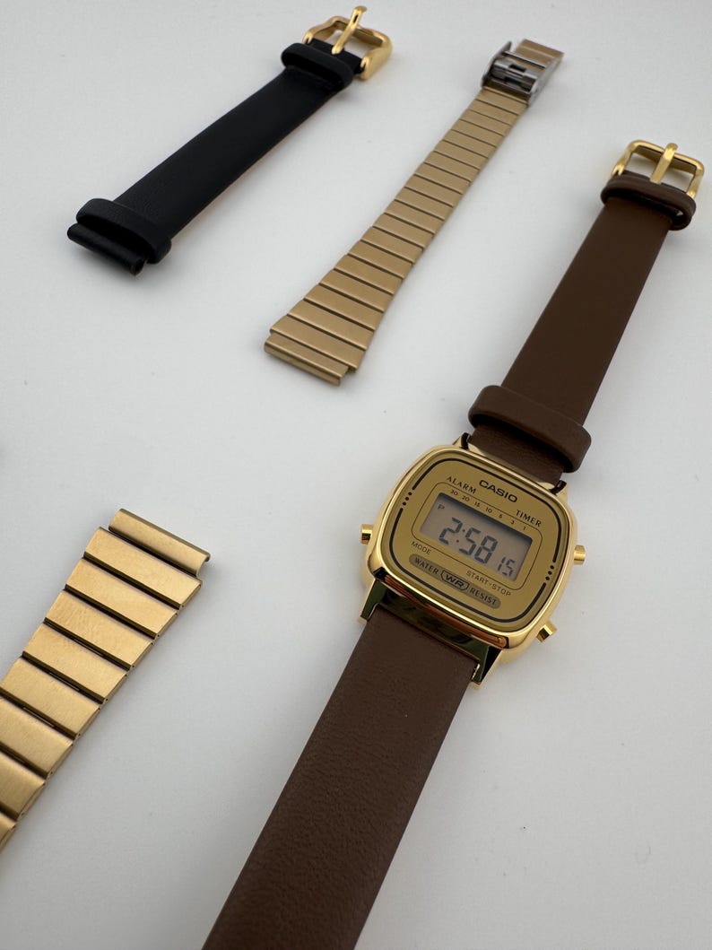 K&ouml;nnte beinhalten: Eine goldfarbene Digitaluhr mit braunem Lederarmband, dazu ein schwarzes Lederarmband und zwei goldfarbene Metallarmb&auml;nder. Das Zifferblatt der Uhr ist rechteckig und zeigt die Uhrzeit an.