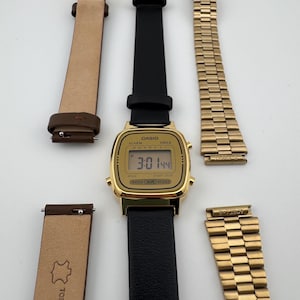 K&ouml;nnte beinhalten: Eine goldfarbene Casio Digitaluhr mit schwarzem Lederarmband wird gezeigt. Zwei zus&auml;tzliche Armb&auml;nder sind enthalten: ein braunes Lederarmband und ein goldenes Metallarmband. Das Zifferblatt zeigt die Uhrzeit 3:01:44 an.
