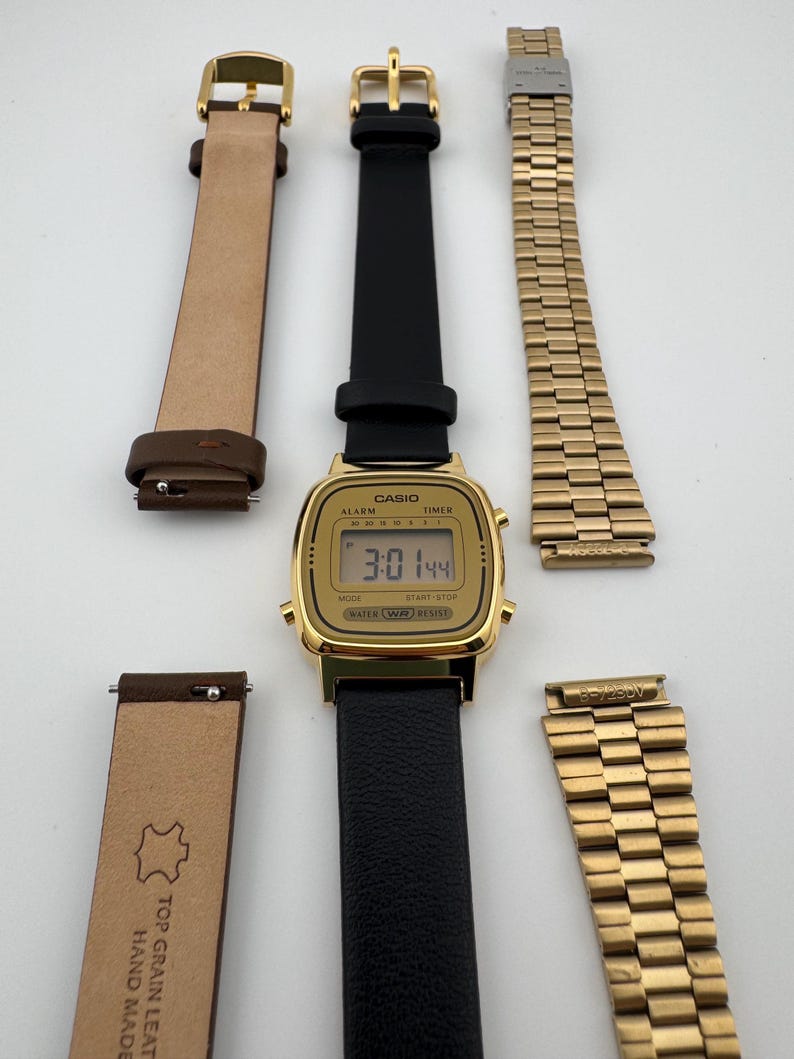 K&ouml;nnte beinhalten: Eine goldfarbene Casio Digitaluhr mit schwarzem Lederarmband wird gezeigt. Zwei zus&auml;tzliche Armb&auml;nder sind enthalten: ein braunes Lederarmband und ein goldenes Metallarmband. Das Zifferblatt zeigt die Uhrzeit 3:01:44 an.