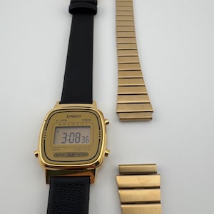 K&ouml;nnte beinhalten: Eine goldfarbene Casio Digitaluhr mit schwarzem Lederarmband und einem separaten goldenen Metallarmband. Das Zifferblatt zeigt die Uhrzeit 3:08:36 an. Die Uhr hat Kn&ouml;pfe f&uuml;r Alarm, Timer, Modus und Start-Stopp-Funktionen.