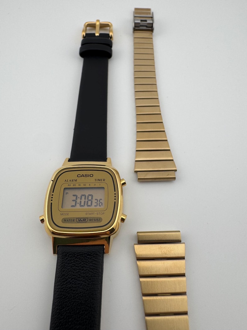 K&ouml;nnte beinhalten: Eine goldfarbene Casio Digitaluhr mit schwarzem Lederarmband und einem separaten goldenen Metallarmband. Das Zifferblatt zeigt die Uhrzeit 3:08:36 an. Die Uhr hat Kn&ouml;pfe f&uuml;r Alarm, Timer, Modus und Start-Stopp-Funktionen.