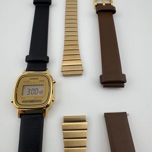 K&ouml;nnte beinhalten: Eine goldfarbene Casio Digitaluhr mit schwarzem Lederarmband, die die Uhrzeit 3:00:48 anzeigt. Zus&auml;tzliche Armb&auml;nder in Gold und braunem Leder sind ebenfalls abgebildet, zusammen mit goldenen Schnallen. Die Uhr ist wasserdicht.