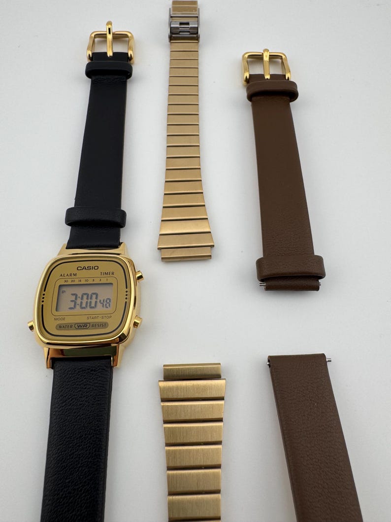 K&ouml;nnte beinhalten: Eine goldfarbene Casio Digitaluhr mit schwarzem Lederarmband, die die Uhrzeit 3:00:48 anzeigt. Zus&auml;tzliche Armb&auml;nder in Gold und braunem Leder sind ebenfalls abgebildet, zusammen mit goldenen Schnallen. Die Uhr ist wasserdicht.