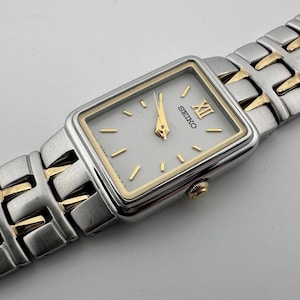 May include: A rectangular Seiko watch with a silver face and gold-tone hands and markers. The watch has a silver and gold-tone band. The watch face displays the Seiko logo and the Roman numeral XII.