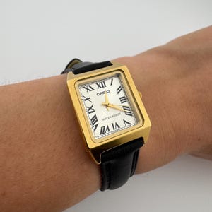 May include: Gold-tone rectangular Casio watch with a white face and black Roman numerals. The watch has a black leather band and the word "CASIO" is printed on the face. The watch is water resistant.