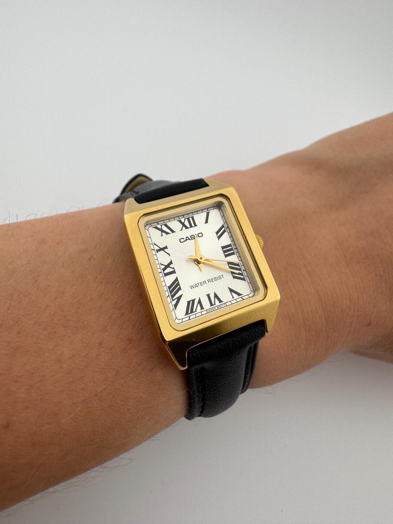 May include: Gold-tone rectangular Casio watch with a white face and black Roman numerals. The watch has a black leather band and the word "CASIO" is printed on the face. The watch is water resistant.