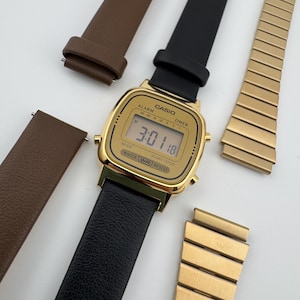 K&ouml;nnte beinhalten: Eine goldfarbene Casio Digitaluhr mit schwarzem Lederarmband zeigt die Uhrzeit 3:01:18 an. Zus&auml;tzliche Armb&auml;nder aus braunem Leder und goldfarbenem Metall sind ebenfalls vorhanden. Das Zifferblatt enth&auml;lt den Text "CASIO", "ALARM", "TIMER", "WATER RESIST".