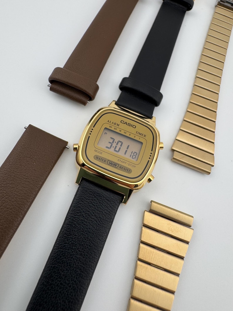 K&ouml;nnte beinhalten: Eine goldfarbene Casio Digitaluhr mit schwarzem Lederarmband zeigt die Uhrzeit 3:01:18 an. Zus&auml;tzliche Armb&auml;nder aus braunem Leder und goldfarbenem Metall sind ebenfalls vorhanden. Das Zifferblatt enth&auml;lt den Text "CASIO", "ALARM", "TIMER", "WATER RESIST".