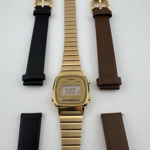 K&ouml;nnte beinhalten: Eine goldfarbene digitale Casio-Uhr mit passendem, ausziehbarem Armband und zwei S&auml;tzen Uhrenarmb&auml;ndern aus schwarzem und braunem Leder. Das Zifferblatt zeigt die Uhrzeit 3:00:12 an. Die Uhr hat ein rechteckiges Zifferblatt und eine goldene Schnalle.