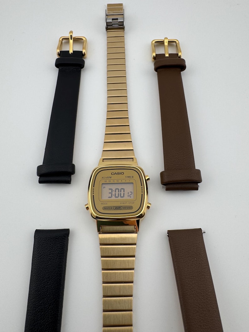 K&ouml;nnte beinhalten: Eine goldfarbene digitale Casio-Uhr mit passendem, ausziehbarem Armband und zwei S&auml;tzen Uhrenarmb&auml;ndern aus schwarzem und braunem Leder. Das Zifferblatt zeigt die Uhrzeit 3:00:12 an. Die Uhr hat ein rechteckiges Zifferblatt und eine goldene Schnalle.