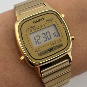 K&ouml;nnte beinhalten: Goldfarbene Casio Digitaluhr mit rechteckigem Zifferblatt und passendem Armband. Die Uhr zeigt die Zeit 12:30:16 an. Funktionen: Alarm, Timer, Modus, Start-Stopp und Wasserdichtigkeit. Ein klassisches und stilvolles Accessoire.