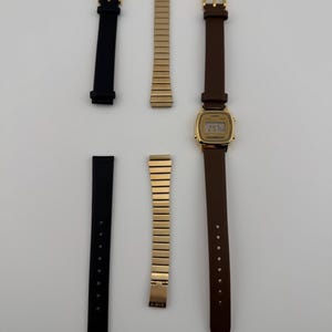 K&ouml;nnte beinhalten: Eine goldfarbene Digitaluhr mit braunem Lederarmband und zwei schwarzen Armb&auml;ndern. Ebenfalls enthalten sind zwei goldfarbene Metallarmb&auml;nder. Die Uhr zeigt die Uhrzeit an. Die Artikel sind auf einer wei&szlig;en Oberfl&auml;che angeordnet.