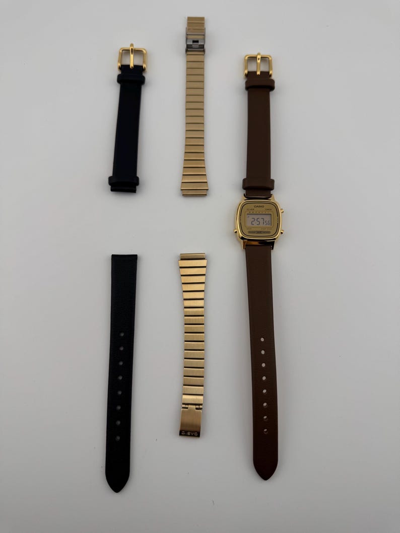 K&ouml;nnte beinhalten: Eine goldfarbene Digitaluhr mit braunem Lederarmband und zwei schwarzen Armb&auml;ndern. Ebenfalls enthalten sind zwei goldfarbene Metallarmb&auml;nder. Die Uhr zeigt die Uhrzeit an. Die Artikel sind auf einer wei&szlig;en Oberfl&auml;che angeordnet.