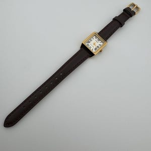 May include: A rectangular gold-toned wristwatch with a white face and Roman numeral hour markers. The watch has a dark brown leather band with a gold-colored buckle. The watch is angled diagonally across a white surface.