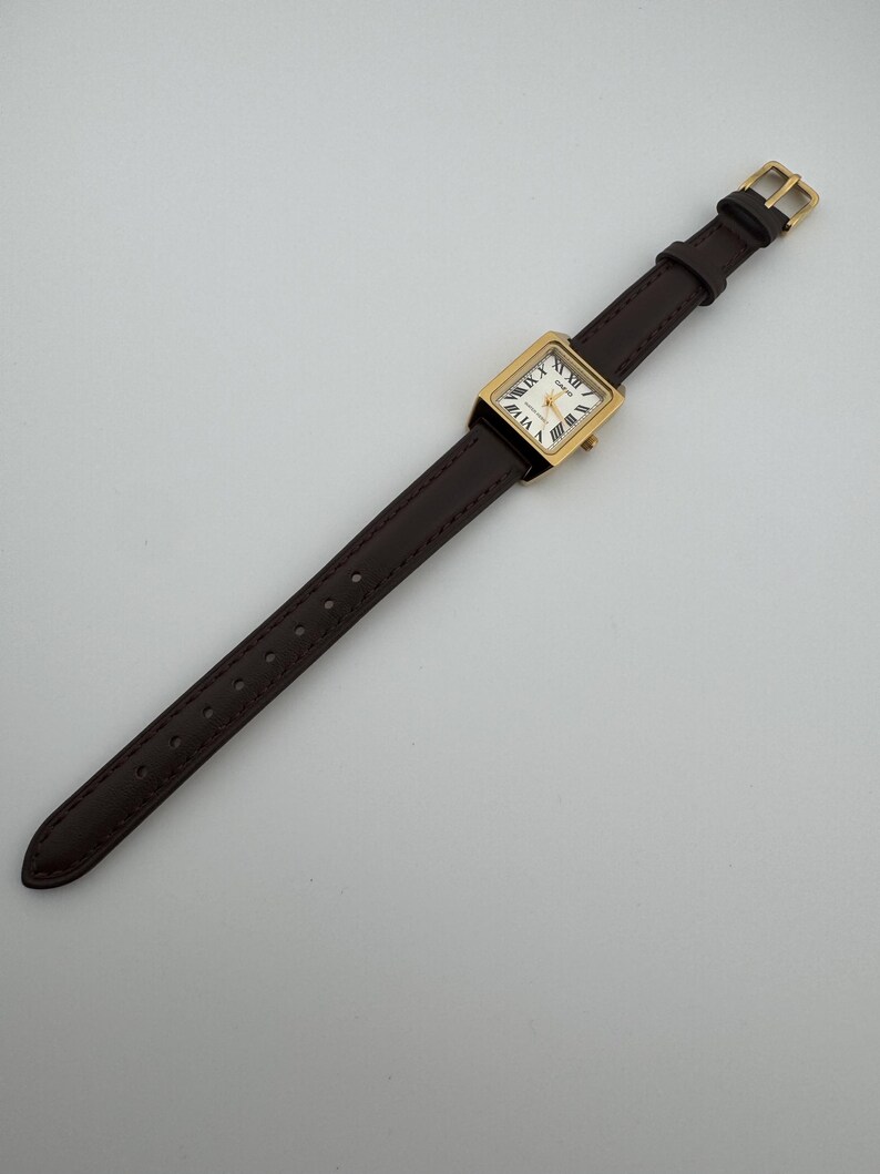 May include: A rectangular gold-toned wristwatch with a white face and Roman numeral hour markers. The watch has a dark brown leather band with a gold-colored buckle. The watch is angled diagonally across a white surface.