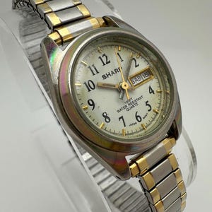 May include: A silver and gold-toned SHARP quartz watch with a white face. The watch features black numerals, gold hands, and a day/date display. The watch band is a combination of silver and gold links. The watch is water resistant.