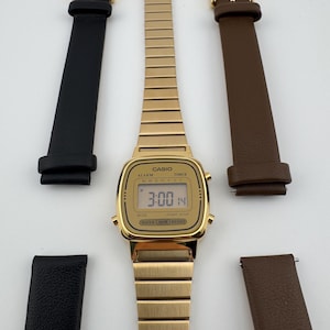 K&ouml;nnte beinhalten: Eine goldfarbene Casio Digitaluhr mit passendem goldenen Armband, zusammen mit zwei Lederarmb&auml;ndern, einem schwarzen und einem braunen. Das Zifferblatt zeigt die Uhrzeit 3:00 und das Datum 14 an.