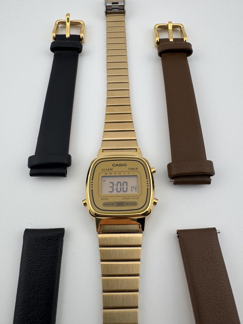 K&ouml;nnte beinhalten: Eine goldfarbene Casio Digitaluhr mit passendem goldenen Armband, zusammen mit zwei Lederarmb&auml;ndern, einem schwarzen und einem braunen. Das Zifferblatt zeigt die Uhrzeit 3:00 und das Datum 14 an.