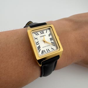 May include: A gold-toned rectangular Casio watch with a white face and black Roman numerals. The watch has a black leather band and gold hands. The watch face reads "WATER RESIST".