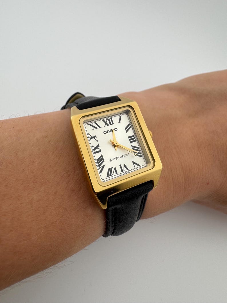 May include: A gold-toned rectangular Casio watch with a white face and black Roman numerals. The watch has a black leather band and gold hands. The watch face reads "WATER RESIST".