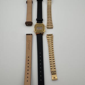 K&ouml;nnte beinhalten: Eine goldfarbene Digitaluhr mit schwarzem Lederarmband und zwei zus&auml;tzlichen Armb&auml;ndern. Eines ist ein hellbraunes Lederarmband, das andere ein goldfarbenes Metallarmband. Das Zifferblatt der Uhr zeigt die Uhrzeit an.