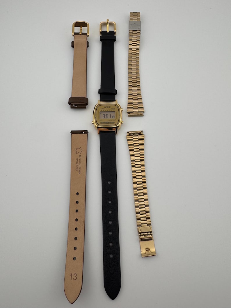 K&ouml;nnte beinhalten: Eine goldfarbene Digitaluhr mit schwarzem Lederarmband und zwei zus&auml;tzlichen Armb&auml;ndern. Eines ist ein hellbraunes Lederarmband, das andere ein goldfarbenes Metallarmband. Das Zifferblatt der Uhr zeigt die Uhrzeit an.