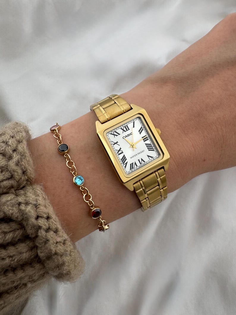 May include: A gold-toned rectangular wristwatch with a white face and Roman numeral hour markers is shown on a wrist. A gold bracelet with colorful gemstones is also visible. The watch band and case are gold-colored.
