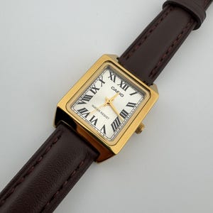 May include: A gold-toned rectangular Casio watch with a white face and black Roman numerals. The watch features a brown leather strap. The watch face reads "CASIO" and "WATER RESIST".