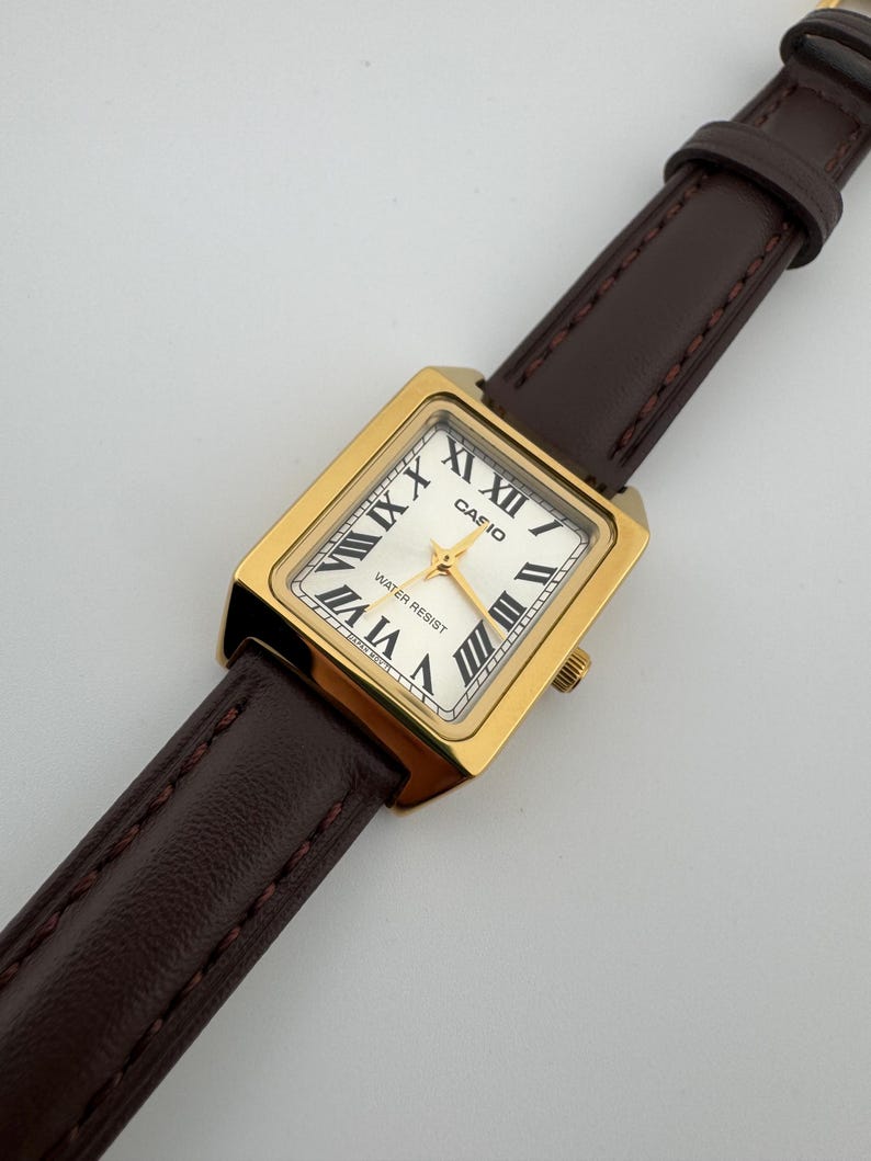 May include: A gold-toned rectangular Casio watch with a white face and black Roman numerals. The watch features a brown leather strap. The watch face reads "CASIO" and "WATER RESIST".