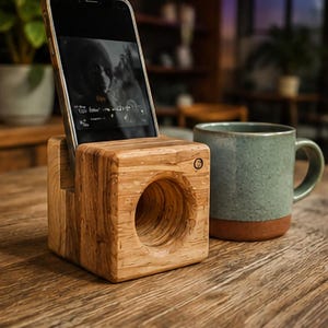 May include: A square wooden smartphone stand featuring a natural grain pattern and a circular sound amplification chamber on the front face. The device holder includes a slot for a phone and a small circular power button icon on the top right corner.