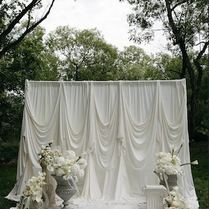 May include: A draped white fabric backdrop for an outdoor event, with cascading folds and a soft, elegant appearance. White floral arrangements in ornate urns and on pedestals add to the romantic setting. The scene is set on a grassy area, framed by trees.