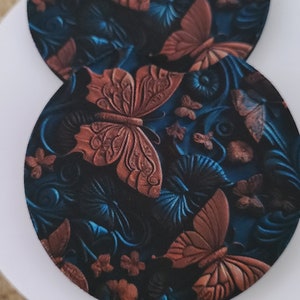 May include: Two round coasters with a butterfly design. The coasters feature a dark blue background with embossed rust-colored butterflies and floral accents. The coasters are placed on a white surface.
