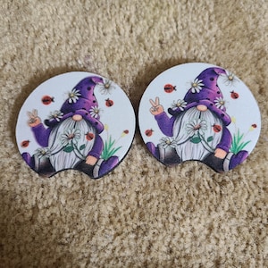 May include: Two round white coasters with a gnome design. Each gnome is wearing a purple hat with white flowers and holding a white flower. The gnomes are surrounded by white flowers and red ladybugs.