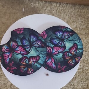 May include: Two round coasters with a dark green background and a colorful butterfly design. The butterflies are pink, purple, and blue with black outlines.