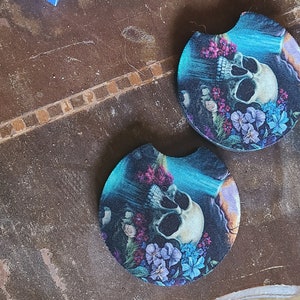 May include: Two round, blue and black coasters with a skull and floral design. The coasters feature a white skull with a blue background and pink and purple flowers.