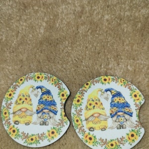 May include: Two round, white coasters with a design of two gnomes wearing yellow and blue hats. The gnomes are surrounded by sunflowers and green leaves.