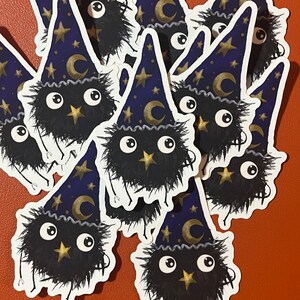 May include: A collection of black fuzzy wizard stickers with gold stars and a crescent moon on their hats. The stickers are on a brown surface.