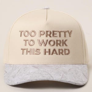 May include: Cream-colored baseball cap with the embroidered text "TOO PRETTY TO WORK THIS HARD." The brim has a marbled gray and white pattern. The cap has a curved brim and a button on top.