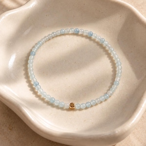 May include: A delicate beaded bracelet featuring small, translucent light blue round beads strung together in a circular pattern, accented by a single, polished gold-toned square bead positioned at the bottom centre of the strand.