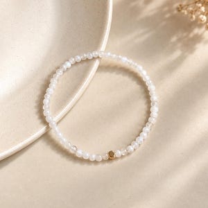 May include: A delicate beaded bracelet featuring small, translucent white round beads arranged in a circular pattern. A single, smooth, metallic gold-toned cube bead is positioned as a focal point among the white beads on the bracelet.