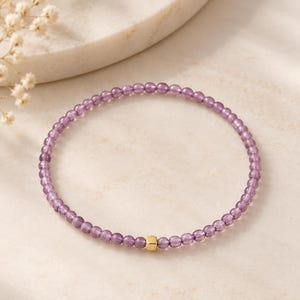 May include: A delicate beaded bracelet featuring small, translucent purple spherical beads strung in a continuous circle, accented by a single, polished gold-toned square bead positioned at the bottom centre of the circular arrangement.
