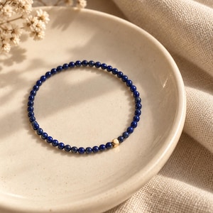 May include: A delicate bracelet composed of small, round, deep blue lapis lazuli beads arranged in a circular pattern, featuring a single, small, polished gold-toned cube bead positioned as a focal point within the continuous string of blue stones.