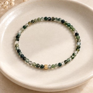 Peut inclure: Un bracelet de perles délicat composé de petites pierres rondes d'agate mousse dans des tons de vert forêt, de blanc translucide et de gris anthracite, agrémenté d'une perle carrée unique en métal doré polie au centre des pierres naturelles.