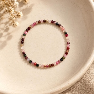 May include: A delicate beaded bracelet featuring small, round, multi-coloured tourmaline gemstones in shades of pink, red, green, and black, accented by a single small, square, gold-toned metal bead positioned at the bottom of the circular strand.