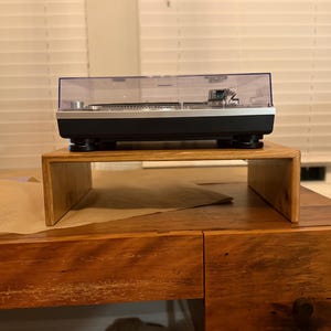 Może przedstawiać: Czarny i srebrny gramofon z przezroczystą osłoną przeciwpyłową stoi na drewnianym stojaku. Gramofon stoi na drewnianym biurku z laptopem w tle.