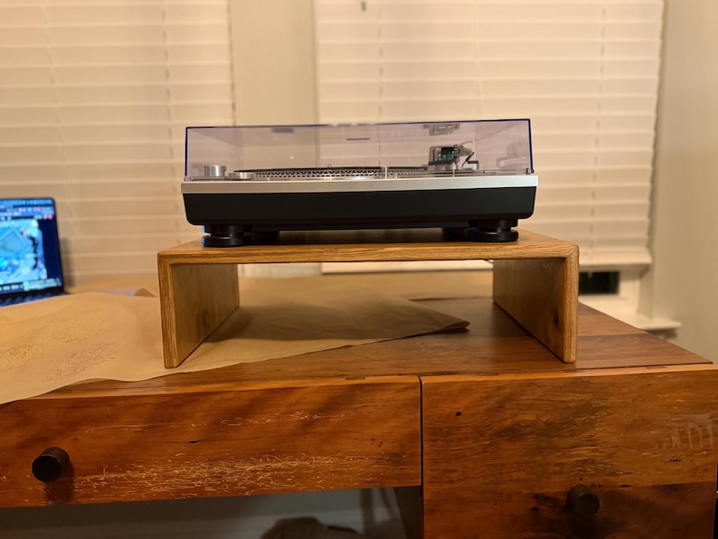 Może przedstawiać: Czarny i srebrny gramofon z przezroczystą osłoną przeciwpyłową stoi na drewnianym stojaku. Gramofon stoi na drewnianym biurku z laptopem w tle.