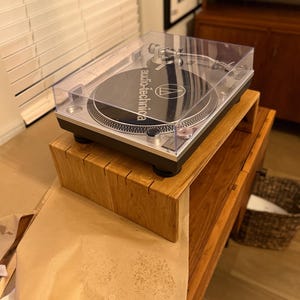 Może przedstawiać: Srebrno-czarny gramofon Audio-Technica z przezroczystą osłoną przeciwpyłową. Gramofon stoi na drewnianym stojaku, kt&oacute;ry umieszczony jest na drewnianej szafce. Gramofon ma czarną płytę.