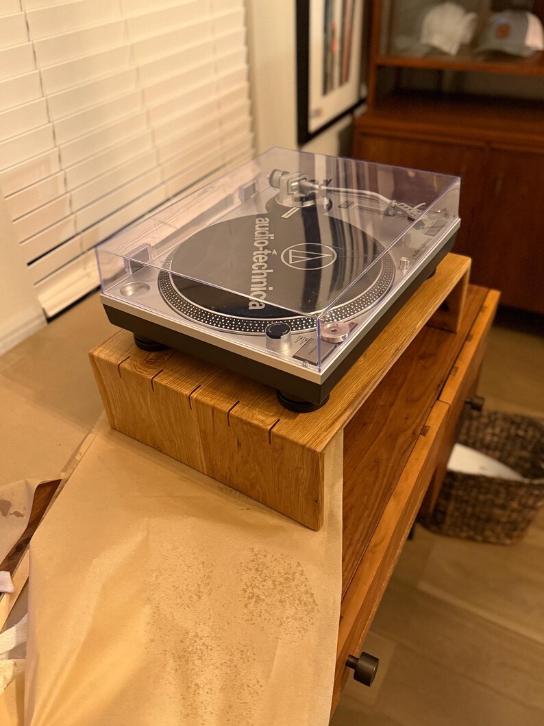 Może przedstawiać: Srebrno-czarny gramofon Audio-Technica z przezroczystą osłoną przeciwpyłową. Gramofon stoi na drewnianym stojaku, kt&oacute;ry umieszczony jest na drewnianej szafce. Gramofon ma czarną płytę.
