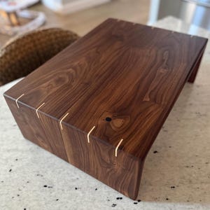 May include: A handcrafted wooden laptop stand. The stand is made of dark walnut wood with visible grain and features angled legs. The corners are reinforced with light-colored accents. The stand is designed to elevate a laptop.